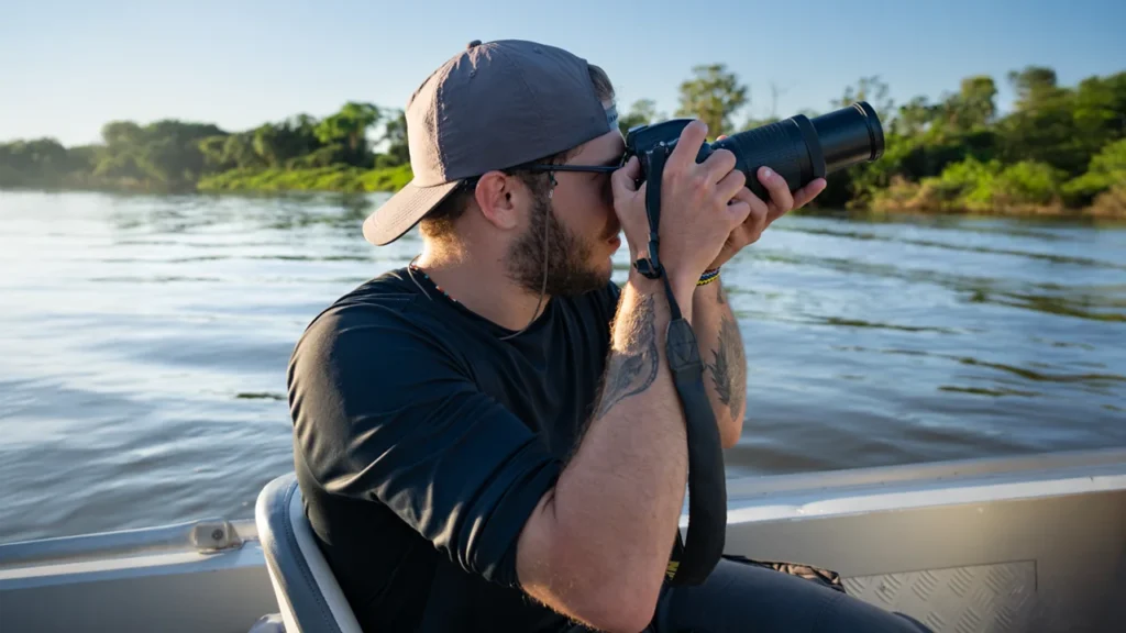Homem com boné e camiseta escura, usando uma câmera DSLR com lente telefoto (zoom) para fotografar a vida selvagem de um barco em movimento no Pantanal.
