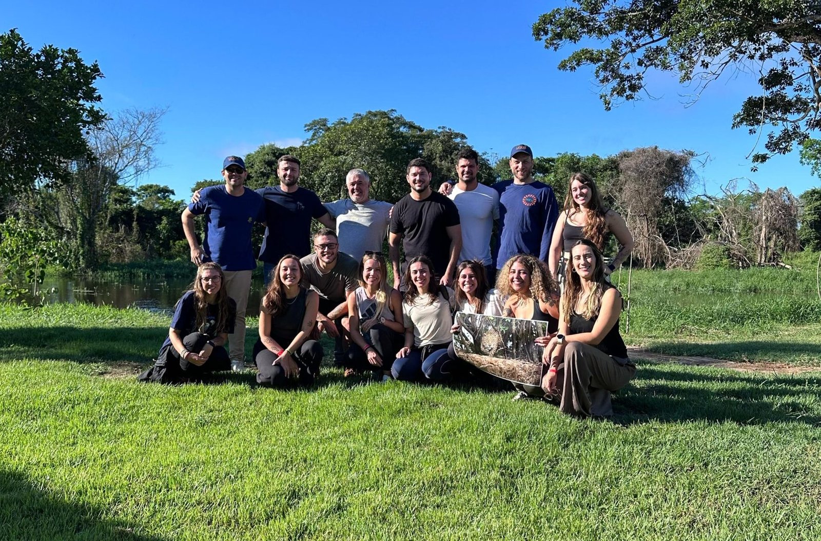 Foto de grupo da equipe Yara Eco Safari e turistas sorrindo em gramado verde no Pantanal, com árvores e lago ao fundo. Representa a hospitalidade e a experiência em grupo nos safaris do Pantanal.