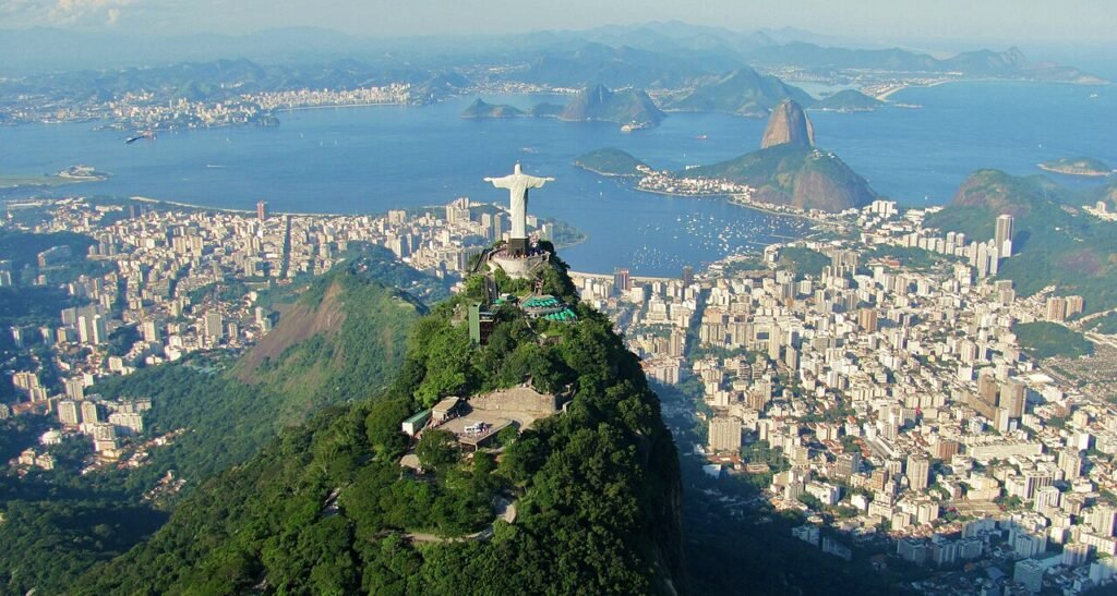 Vista aérea panorâmica do Cristo Redentor no topo do Corcovado, com a cidade do Rio de Janeiro, Pão de Açúcar, Baía de Guanabara e montanhas ao fundo.
