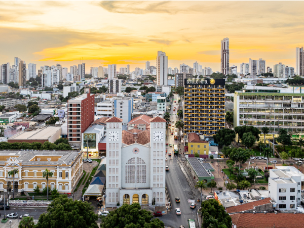 Vista aérea da Catedral Basílica do Senhor Bom Jesus em Cuiabá, com suas duas torres brancas em destaque, cercada por edifícios modernos e ruas movimentadas sob um pôr do sol amarelado.