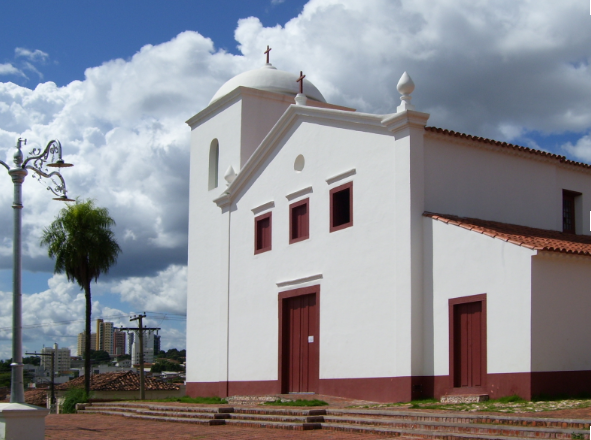 Vista frontal da Igreja de Nossa Senhora do Rosário e São Benedito em Cuiabá, uma construção colonial branca com portas e janelas de madeira marrom, sob um céu azul com nuvens brancas.