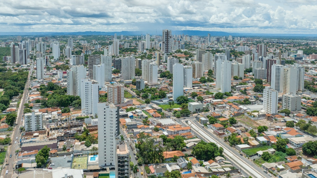 Vista aérea panorùmica de uma cidade brasileira com diversos prédios residenciais brancos de arquitetura moderna, åreas arborizadas entre as ruas e céu nublado ao fundo.