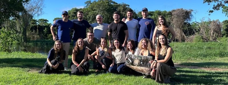 Foto de grupo da equipe Yara Eco Safari e turistas sorrindo em gramado verde no Pantanal, com árvores e lago ao fundo. Representa a hospitalidade e a experiência em grupo nos safaris do Pantanal.