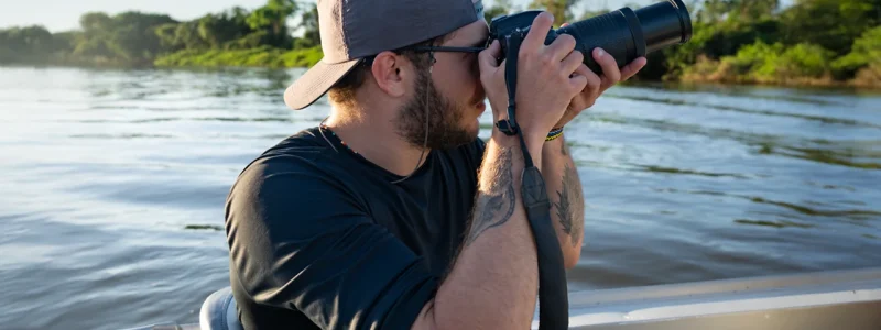 Homem com boné e camiseta escura, usando uma câmera DSLR com lente telefoto (zoom) para fotografar a vida selvagem de um barco em movimento no Pantanal.