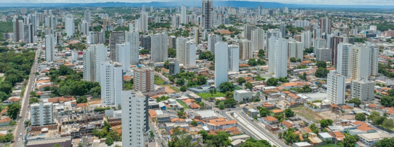 Vista aérea panorâmica de uma cidade brasileira com diversos prédios residenciais brancos de arquitetura moderna, áreas arborizadas entre as ruas e céu nublado ao fundo.